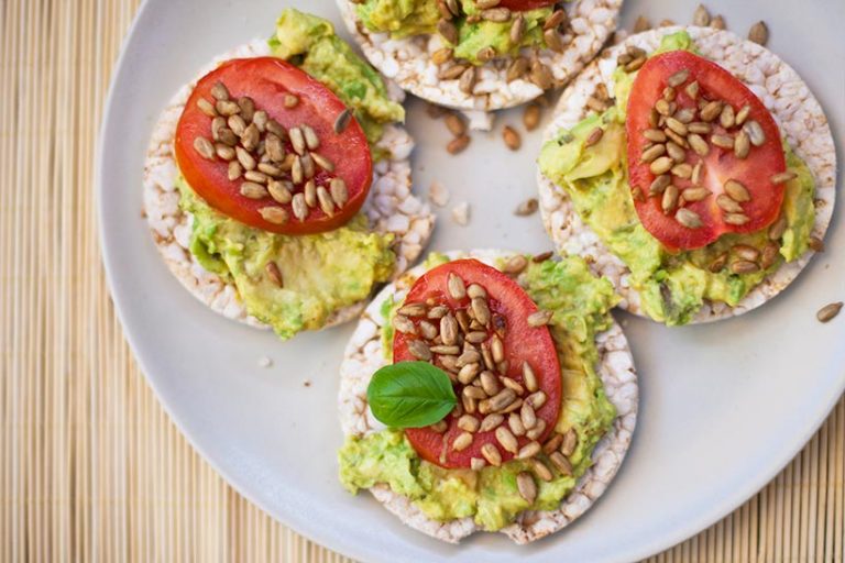 Top rice cakes with avocado, tomato, toasted pine nuts and fresh basil for a healthy snack. 