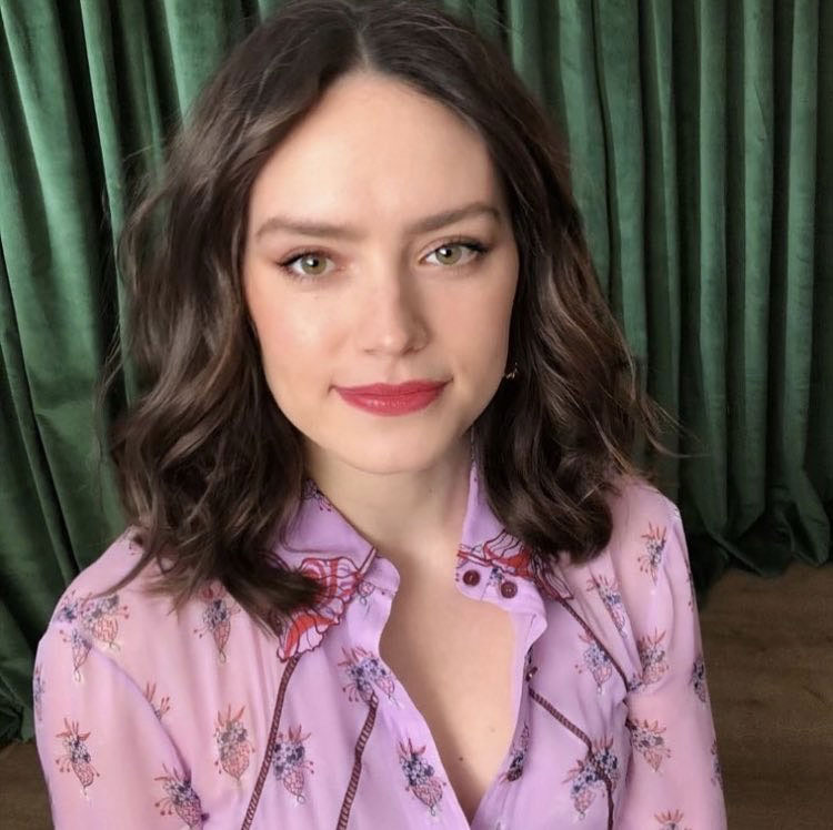 Daisy Ridley wearing a light pink blouse and sitting in front of green curtains 