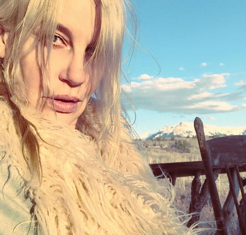 Daryl Hannah wearing a white faux fur coat taking a selfie in front of a mountain range 